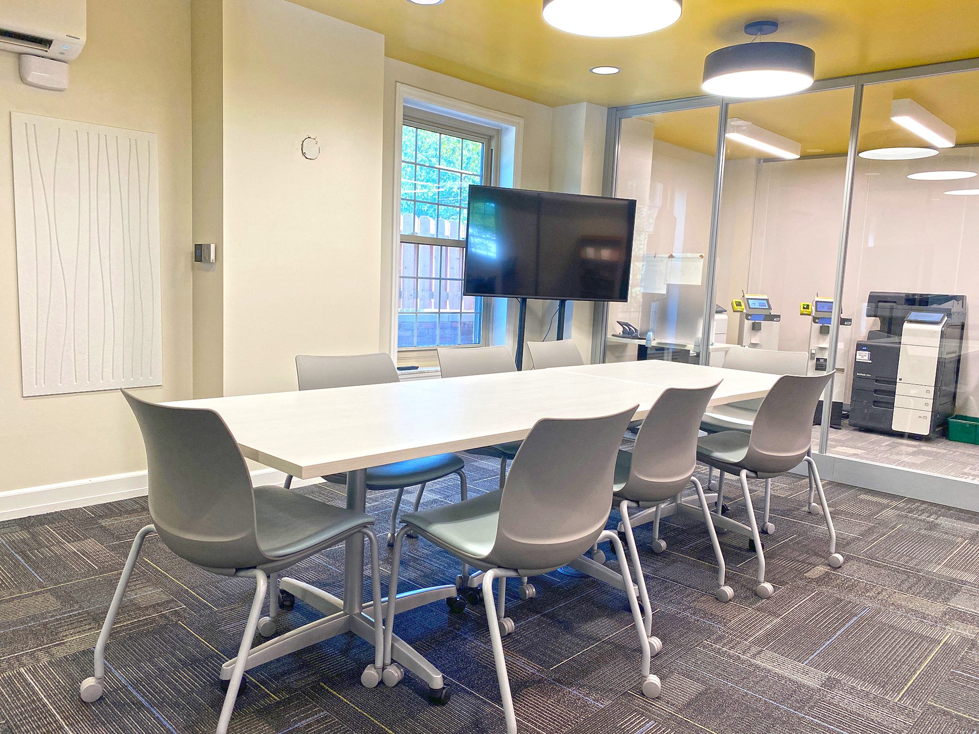 Conference room with a large table, chairs, TV on a stand, yellow ceiling and glass wall.