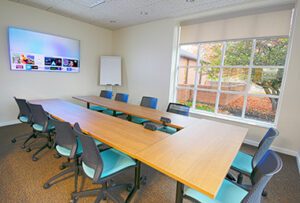 Modern conference room with a large TV, U-shaped table, and aqua chairs, overlooking autumn trees.