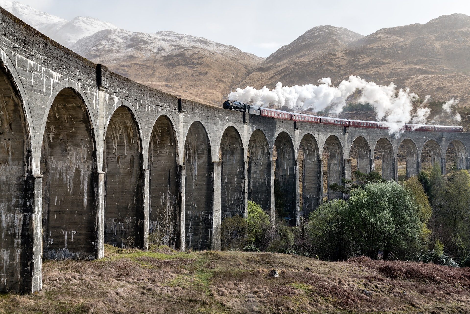 Steam train crossing the Glenfinnan Viaduct in Scotland, with mountains in background and steam billowing from locomotive